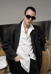 Homme portant des lunettes de soleil noires, un grand manteau gris et une chemise blanche non boutonnée, s'appuyant contre des cartons empilés à l'intérieur.