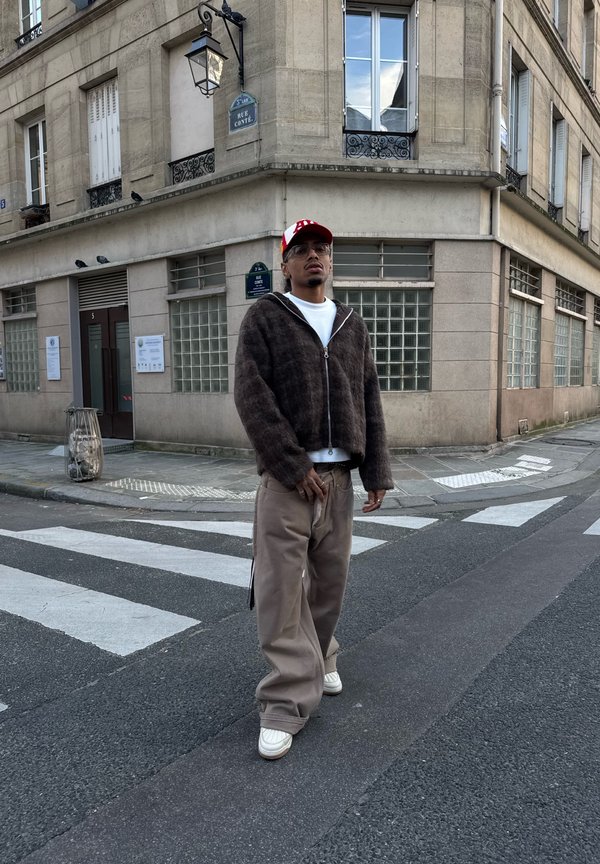 Man wearing a red cap, glasses, dark zip-up jacket, white shirt, and baggy pants standing on a city street crosswalk near a corner building.