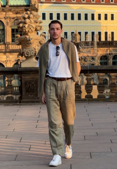 Hombre caminando al aire libre con ropa informal y gafas de sol colgando de la camiseta, edificio histórico y esculturas al fondo.