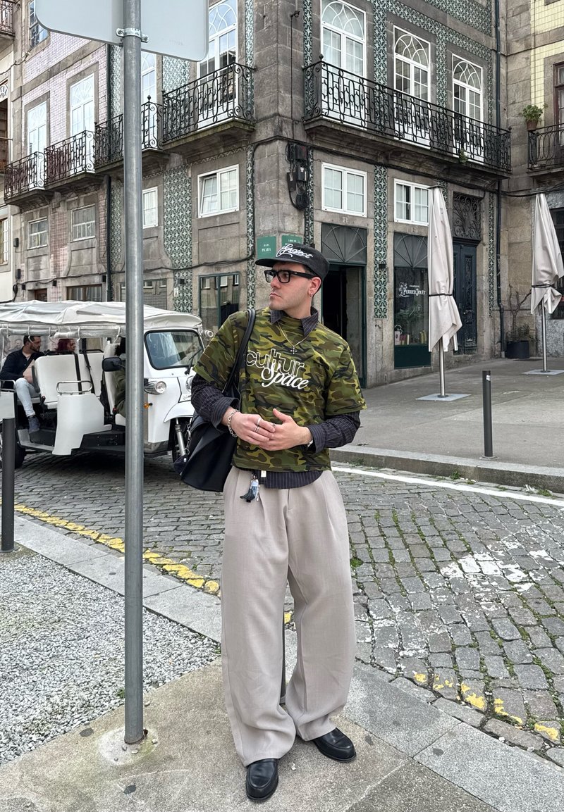 Young man wearing camo shirt, beige pants, and black cap standing on a cobblestone street corner with historic tiled buildings behind.