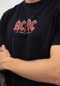 Crna pamučna majica s istrošenim crveno-bijelim grafičkim logotipom "AC/DC" s natpisom "NEKA BUDE ROCK 1977" u središtu.