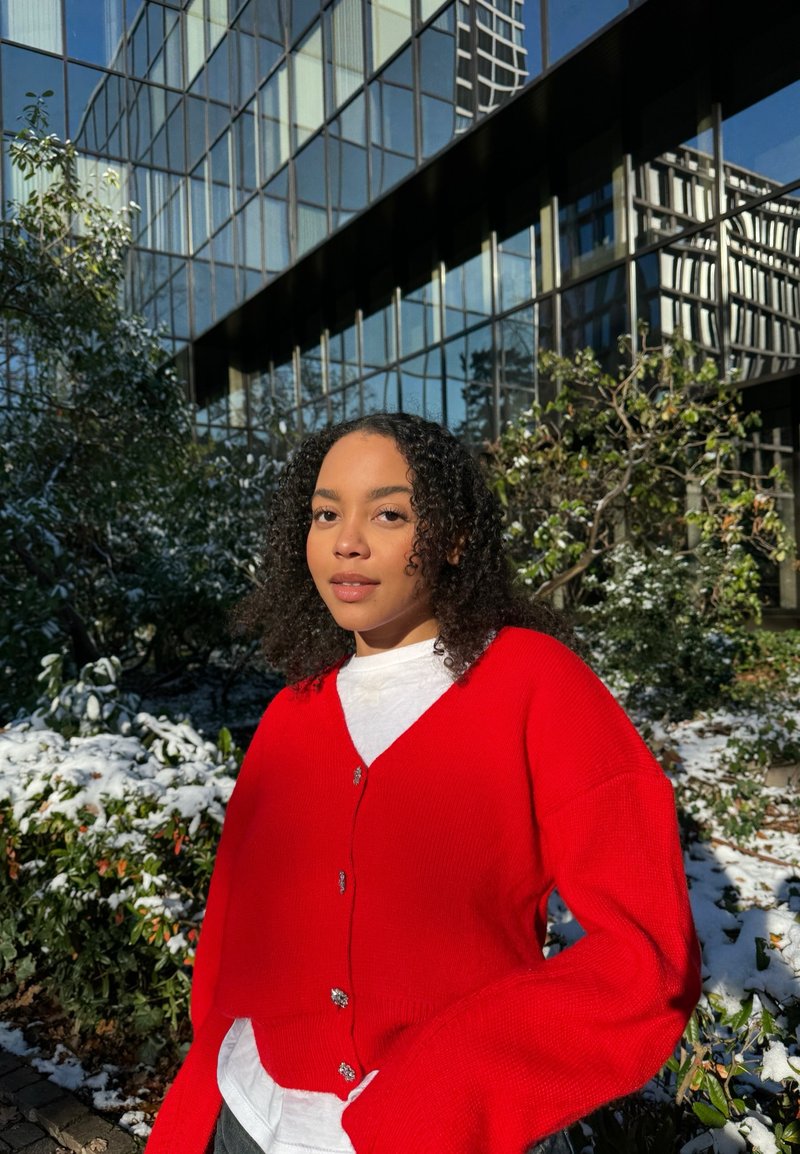 Gilet en tricot rouge avec des manches surdimensionnées et un col en V, agrémenté de boutons argentés, porté sur une chemise blanche, sur fond de neige.