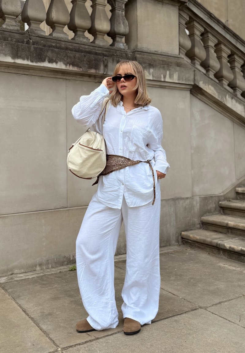 Femme en tenue blanche ample et chaussures marron se tient près d'escaliers en pierre, tenant un sac crème et ajustant des lunettes de soleil noires.