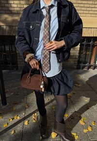 Giacca di denim, camicia azzurro chiaro, cravatta a scacchi, gonna scura, collant neri, stivali alla caviglia marroni e una piccola borsa marrone con dettagli.