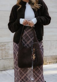 Bolsa hobo de camurça castanha com uma abertura ampla, detalhes de franjas e um pendente. Combinada com uma saia xadrez e um cardigã escuro. Auriculares visíveis.