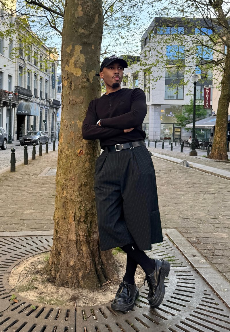 Homme portant une casquette noire, une chemise à manches longues, un pantalon large à fines rayures et des chaussures noires, appuyé contre un arbre dans une rue urbaine avec des bâtiments et des voitures à proximité.