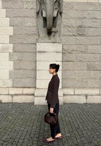 Vrouw in een bruine jas en zwarte broek houdt een donker gevlochten handtas vast, staand zijwaarts op een klinkerpad tegen een stenen muur met een beeld.