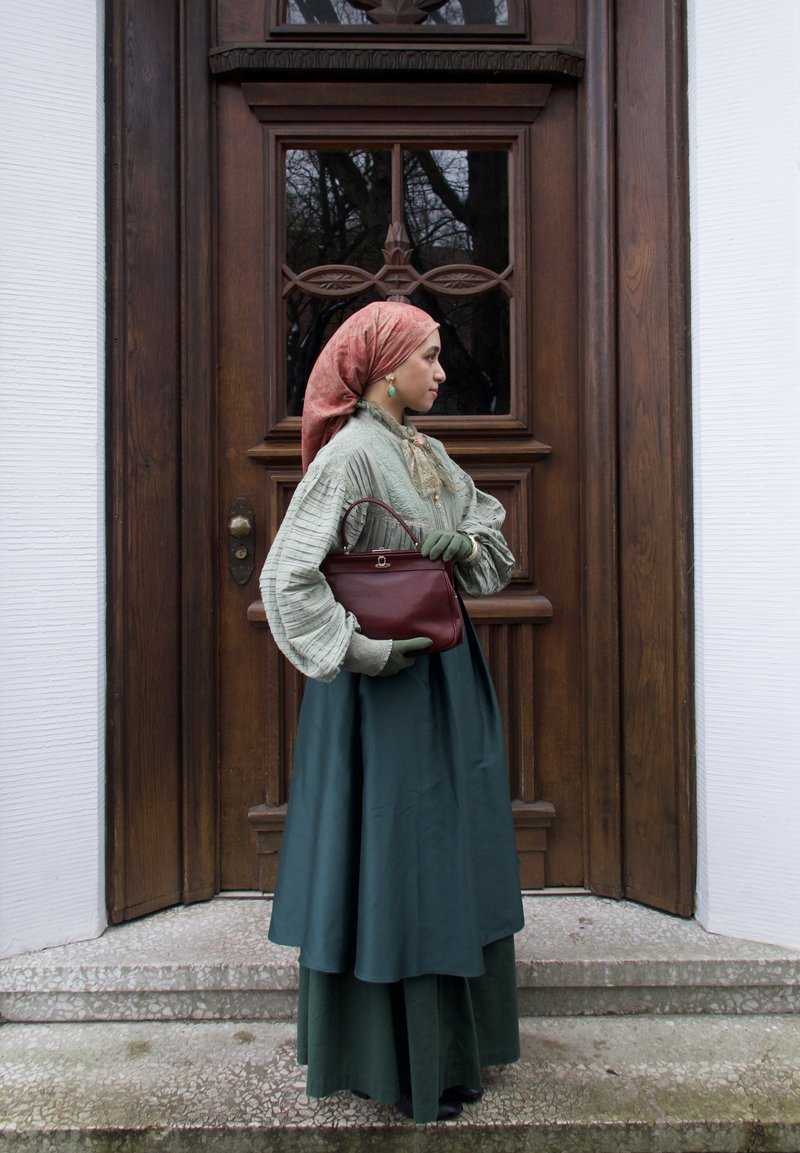 Femme en tenue vintage avec foulard, gants verts et sac à main bordeaux, debout de profil devant une porte en bois.
