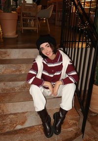 Polo a righe bordeaux e bianche con colletto, pantaloni beige testurizzati e stivali alla caviglia marroni e spessi, seduto su gradini in pietra all'interno.