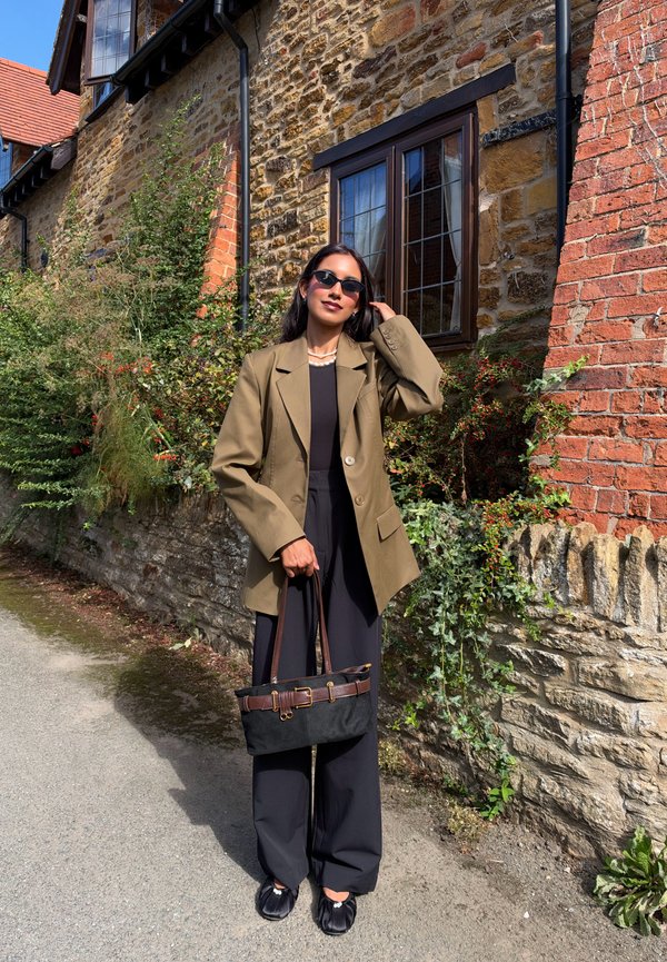 Femme portant des lunettes de soleil et un blazer marron porte un sac à main noir, debout près d'un mur en pierre et d'une fenêtre par une journée ensoleillée.