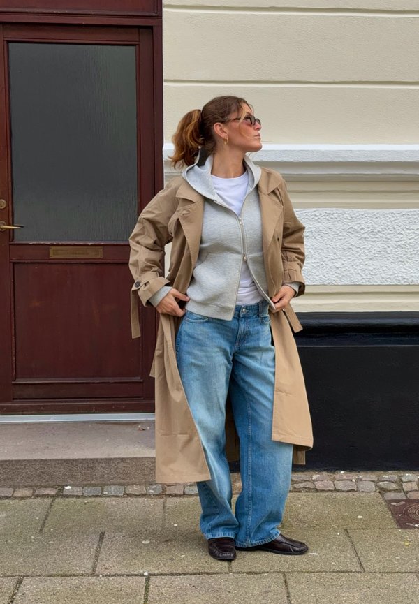Une femme portant des lunettes de soleil, un trench beige, un sweat à capuche gris, une chemise blanche et un jean se tient devant un bâtiment et regarde sur le côté.