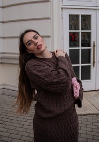 Pull marron tricoté et jupe avec un motif en losanges texturé. Une pochette rose ornée de perles est tenue dans la main droite. En arrière-plan, une porte en verre.