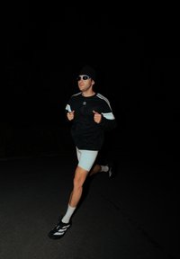 Atletická postava běžící v černé mikině Adidas s bílými pruhy, světlých šortkách, slunečních brýlích a černých teniskách s bílými detaily.