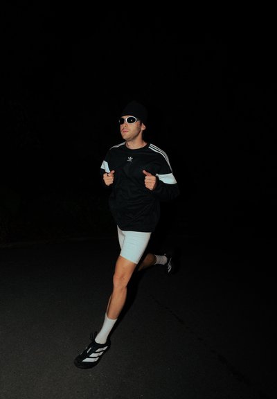 Figura atlética corriendo con una sudadera negra de Adidas con rayas blancas, pantalones cortos de color claro, gafas de sol y zapatillas negras con detalles en blanco.