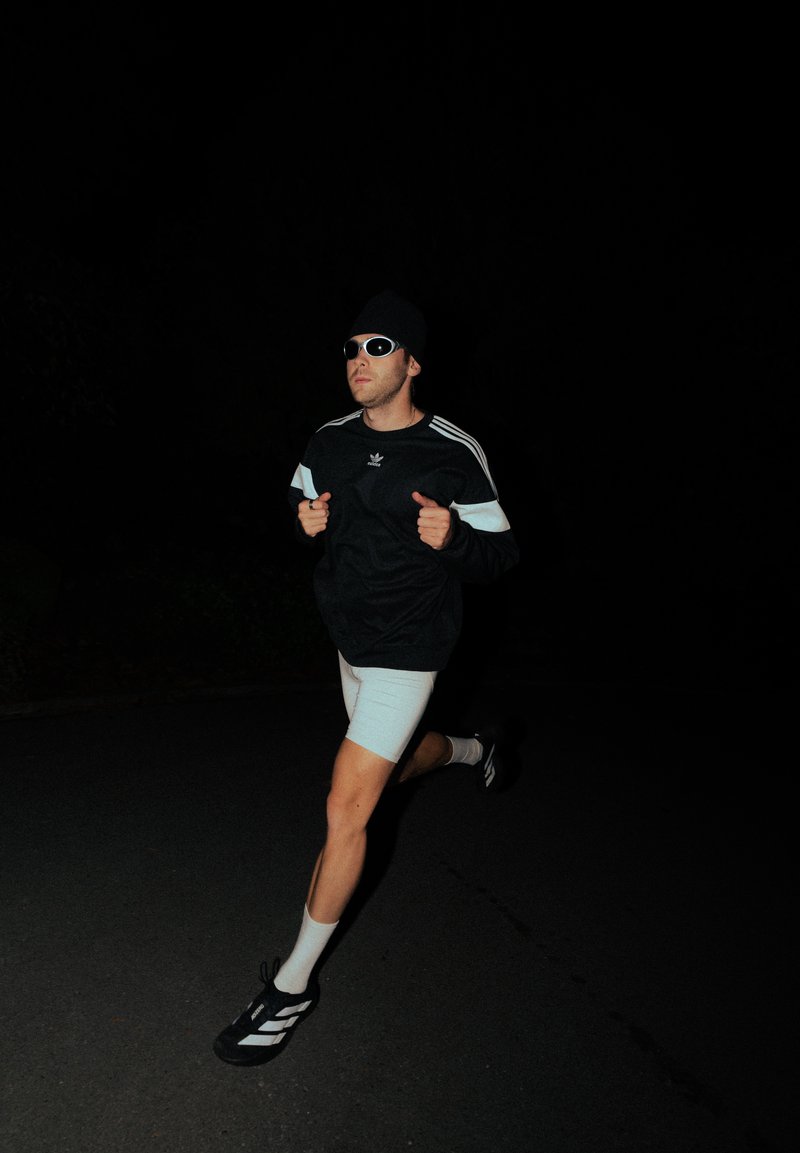 Athletische Figur, die in einem schwarzen Adidas-Sweatshirt mit weißen Streifen, hellen Shorts, einer Sonnenbrille und schwarzen Turnschuhen mit weißen Akzenten läuft.
