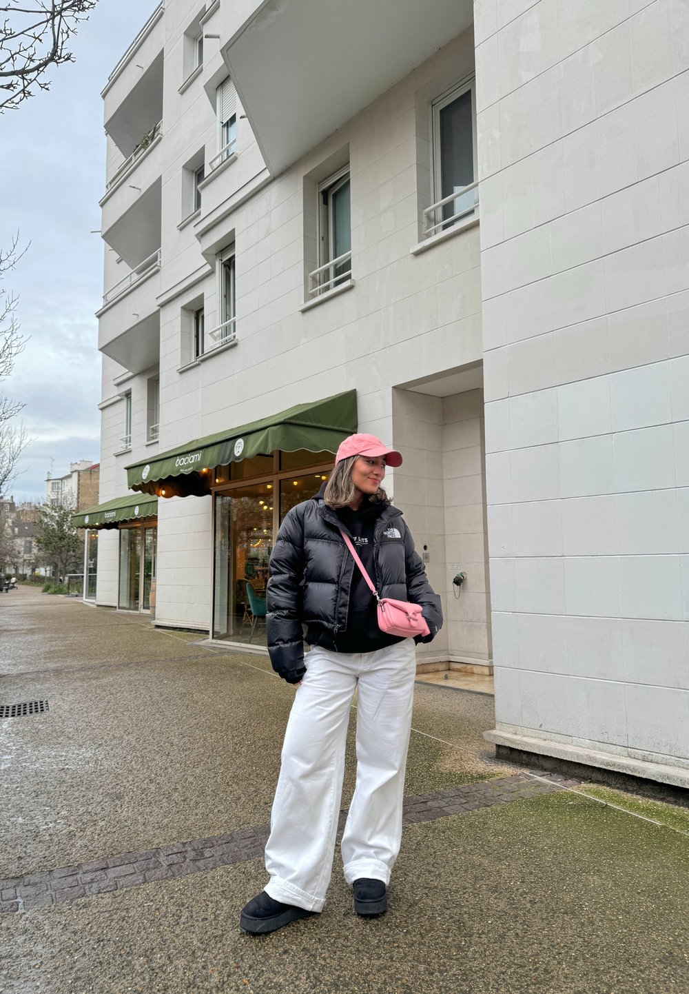 Veste matelassée noire, sweat à capuche noir, pantalon large blanc, sac bandoulière rose et casquette rose ; debout sur un trottoir mouillé près d'un café.