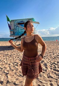 Jeune femme aux cheveux tressés et portant un collier de coquillages, debout sur une plage ensoleillée près d'une tour de surveillance colorée au bord de l'océan.