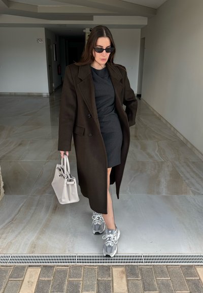 Mujer con abrigo negro y vestido, gafas de sol, zapatillas grises, llevando un bolso blanco, caminando sobre un suelo de baldosas en interiores.