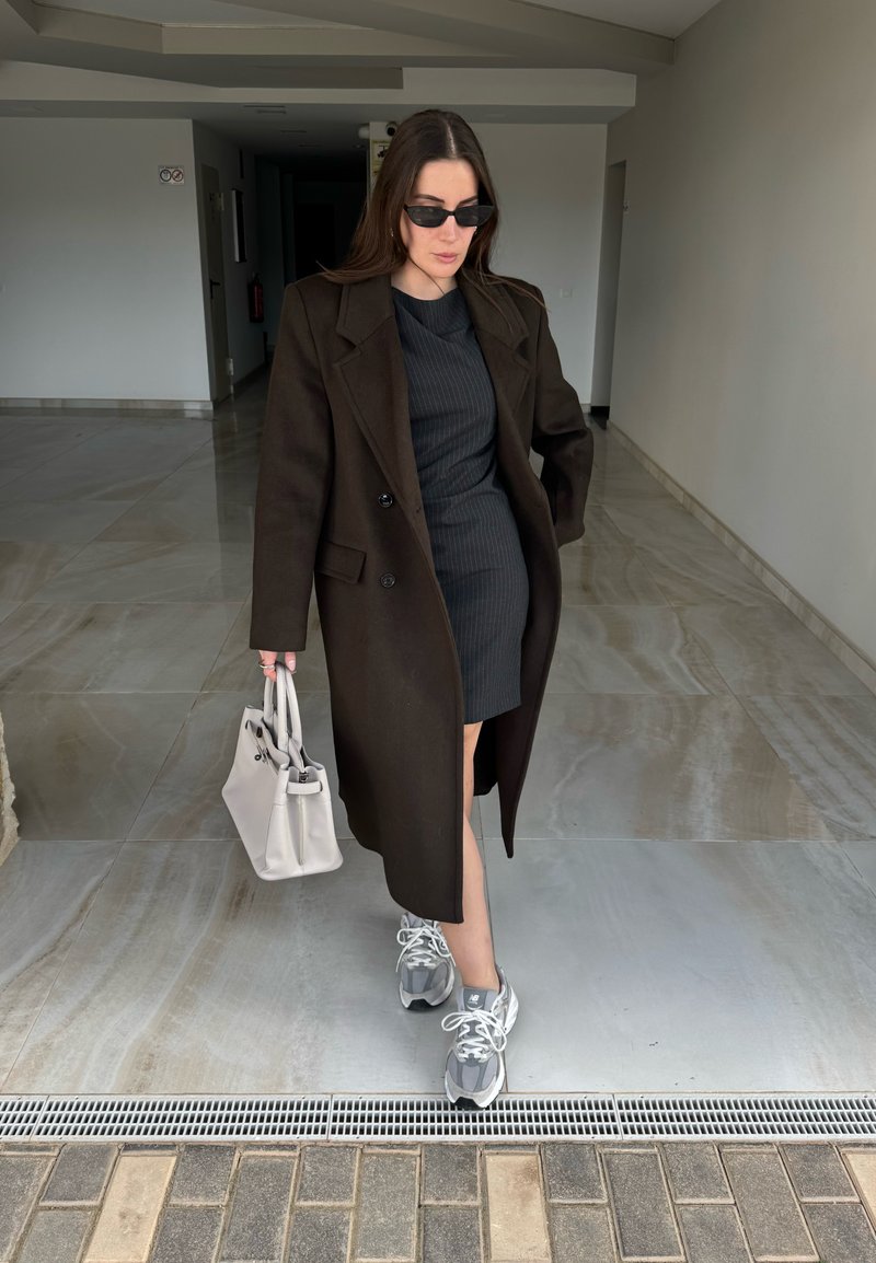 Femme portant un manteau et une robe noirs, des lunettes de soleil, des baskets grises, portant un sac à main blanc, marchant sur un sol carrelé à l'intérieur.