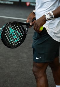 Must värvilise türkiissiniste detailidega aerutennisereket, millel on kuusnurkadega augud, mida hoiab mängija. Taskus on roheline tennispall. Mängija kannab musti lühikesi pükse ja valget särki.