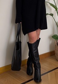 Botas pretas até ao joelho com uma textura de couro suave. Usadas com um vestido de malha preto, combinadas com uma bolsa tote preta. Chão de madeira natural.