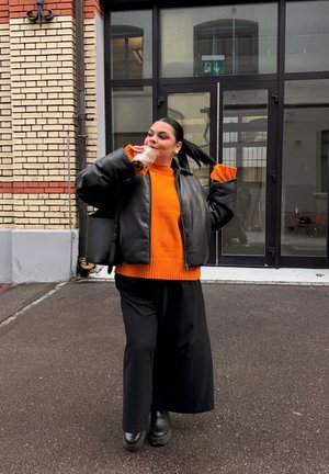 Mujer con chaqueta de cuero negra y suéter naranja bebiendo café con hielo, sujetándose el cabello en una coleta, parada en la calle frente a puertas de cristal.