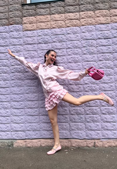 Camisa de satén rosa con mangas abullonadas, falda rosa con volantes en capas, bolso rosa y zapatos planos con adornos, todo sobre un fondo de pared morada.