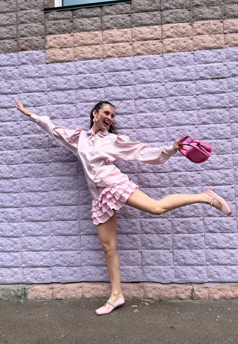 Rosa Satinbluse mit Puffärmeln, geschichteter rosa Volantsrock, rosa Handtasche und flache Schuhe mit Verzierungen, vor einer lila Wand.