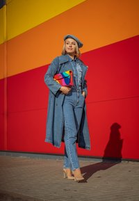 Τζιν καμπαρντίνα πάνω από τζιν πουκάμισο και τζιν παντελόνι, συνδυασμένη με μπεζ τακούνια και μια πολύχρωμη τσάντα. Το φόντο έχει έντονες κίτρινες, πορτοκαλί και κόκκινες ρίγες.