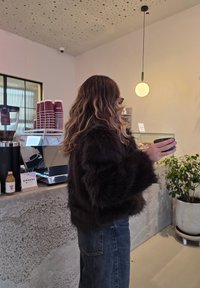 Barna műszőrméből készült kabát bő ujjakkal, sötét farmernadrággal párosítva. A jelenet egy minimalista kávézó belsejét ábrázolja, ahol felszerelések és növények láthatók.