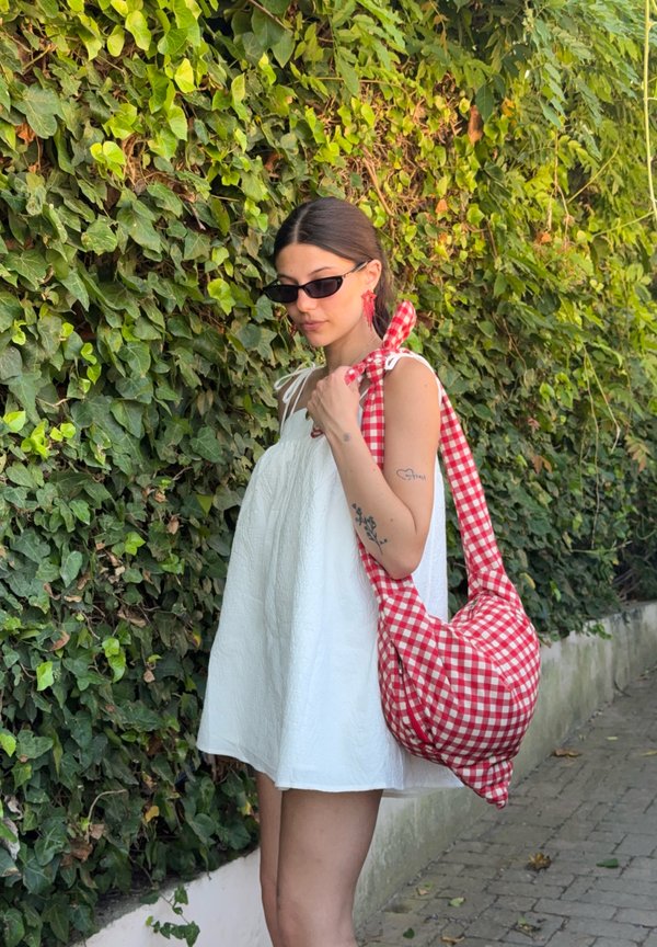 Giovane donna con vestito bianco e occhiali da sole scuri, con una borsa a quadri rossa e bianca sulla spalla, in piedi accanto a un muro verde ricoperto di foglie.