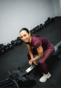Concept2 rowing machine with black frame, grey seat, and footrests. Person in burgundy sportswear and white trainers, smiling. Dumbbells in the background.