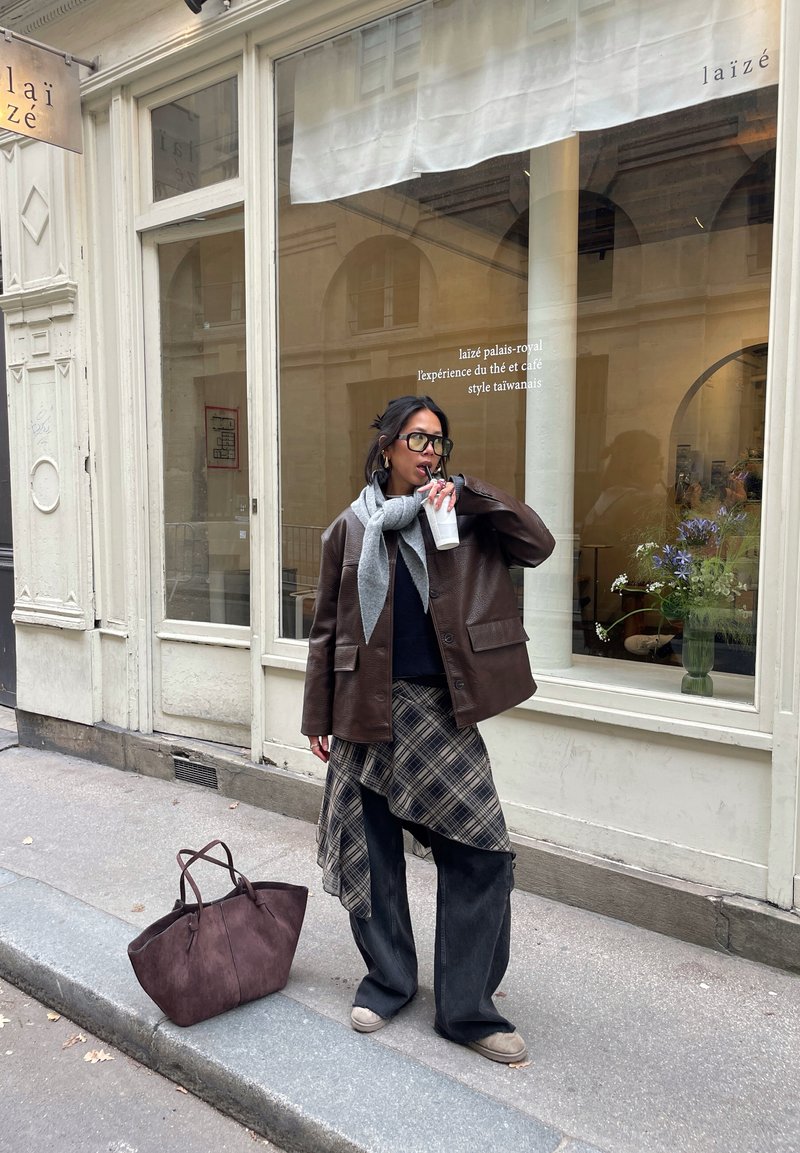 Veste en cuir marron sur un pull sombre, écharpe grise, jupe à carreaux et jean large. Sac cabas marron et chaussures claires. Fenêtre de café en arrière-plan.