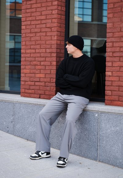 Suéter de punto negro, pantalones grises y zapatillas en blanco y negro. Sentado contra una pared de ladrillo rojo, mostrando un look urbano casual.