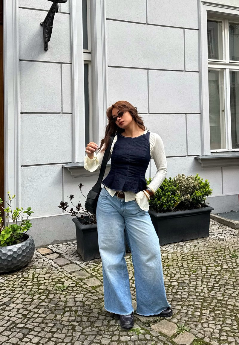 Femme portant un haut noir sans manches sur une chemise blanche à manches longues, un jean large bleu, des chaussures noires, des lunettes de soleil et un sac à bandoulière noir, debout sur des pavés.