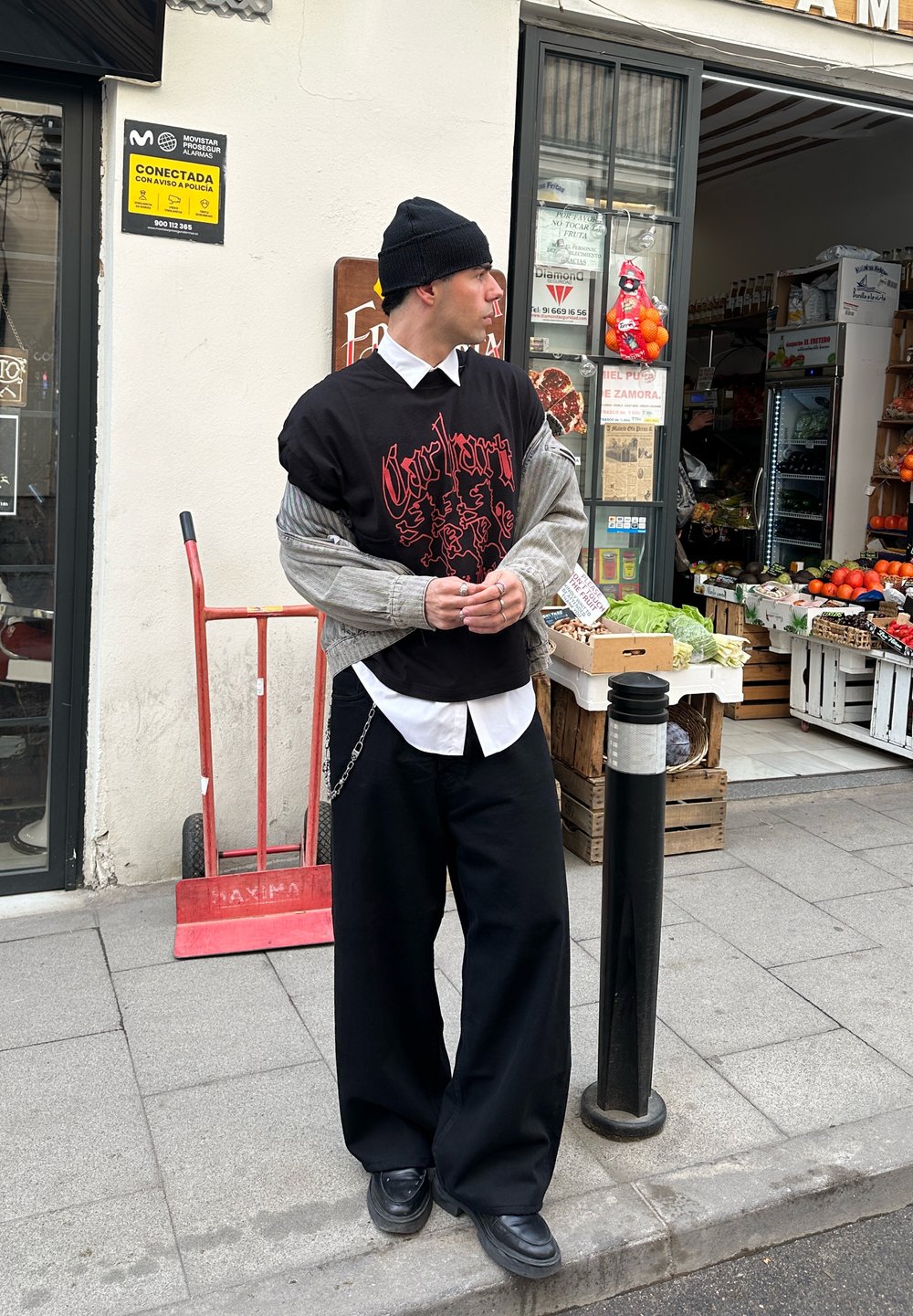 T-shirt noire oversize avec motifs rouges superposée sur une chemise blanche boutonnée, associée à un pantalon large noir et un cardigan gris. Bonnet noir porté.
