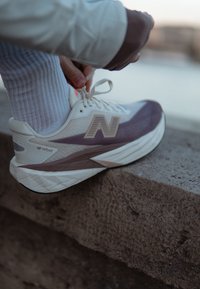 Personne attachant une chaussure de course New Balance blanche et violette en étant assise sur un rebord en pierre en plein air.