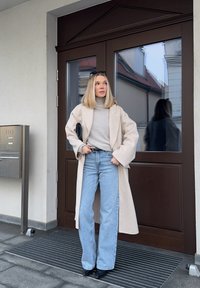 Jeune femme en manteau long beige, col roulé gris clair, jeans bleus et bottes noires, debout avec assurance près d'une porte brun foncé avec des panneaux de verre réfléchissants.