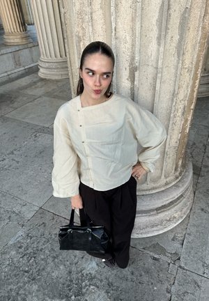 Mujer con el cabello peinado hacia atrás, vestida con una blusa crema y pantalones negros, sosteniendo un bolso negro, de pie junto a grandes columnas de piedra.