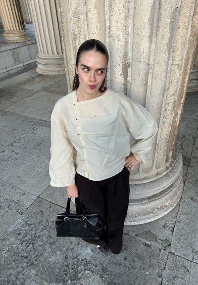 Mujer con el cabello peinado hacia atrás, vestida con una blusa crema y pantalones negros, sosteniendo un bolso negro, de pie junto a grandes columnas de piedra.