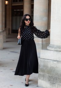 Gonna nera a pieghe abbinata a una blusa nera con pois. Gli accessori includono una piccola borsa nera e occhiali da sole oversize. Tessuto liscio e fluido.