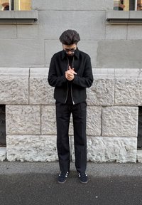 Homme en veste noire, pantalon et baskets, debout sur le trottoir, regardant vers le bas tout en joignant ses mains devant un mur de pierre.