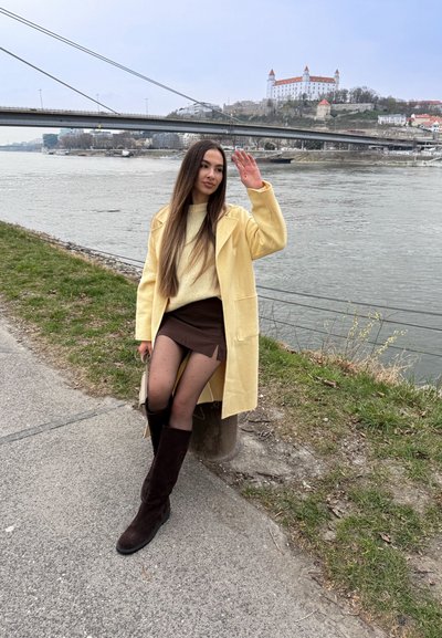 Mujer con abrigo amarillo y botas marrones sentada cerca del río, saludando con puente y castillo al fondo en un día nublado.
