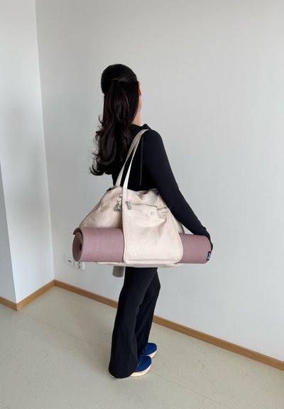 Bolsa de yoga beige con un bolsillo con cremallera, sujetando una esterilla de yoga morada enrollada. Parte superior y pantalones de manga larga negros, zapatos azules, de pie en un interior.