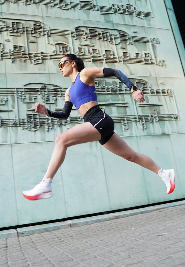 Athletische Frau in blauem Top und schwarzen Shorts springt in der Luft nach vorne vor einer Wand mit großen Musiknotenskulpturen.