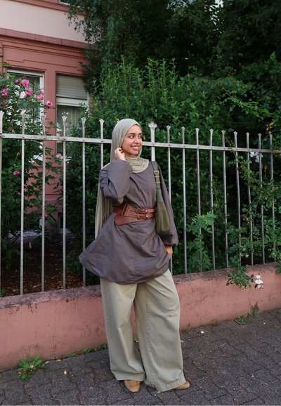 Camisa marrón de manga larga con cintura ceñida, pantalones de pierna ancha verde claro y una bufanda color topo, combinados con zapatos de mimbre beige, todo ello sobre un fondo de vegetación.