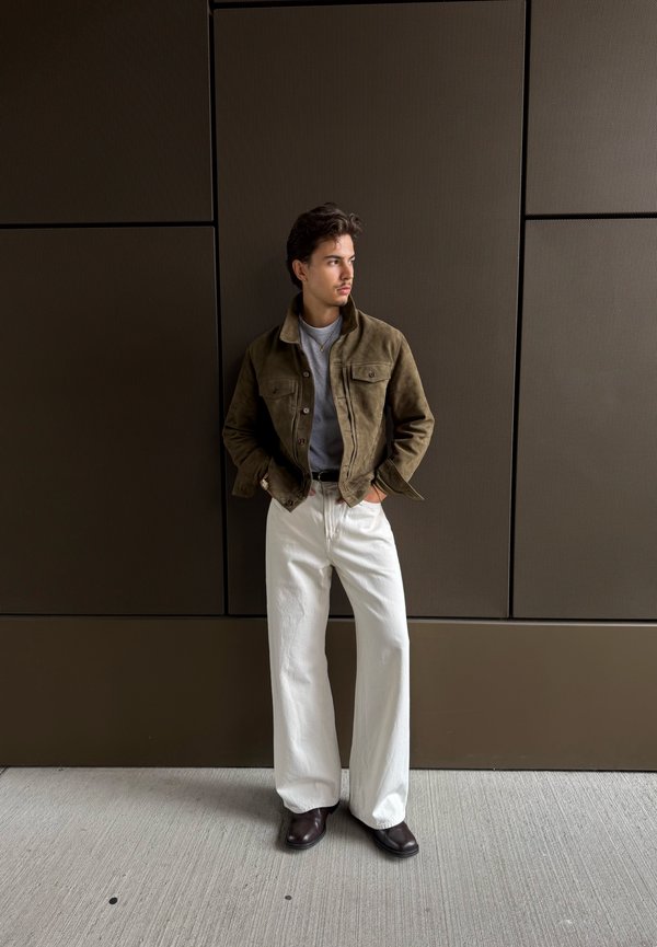 Joven con chaqueta marrón, camisa gris, pantalones blancos de pierna ancha y zapatos oscuros, de pie contra una pared oscura con paneles, con las manos en los bolsillos.
