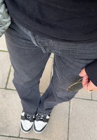 Črne denim hlače z rahlo zbledelim videzom, oblečene z črno-belimi supergami. Sončna očala v roki, postavljena na kamniti površini.