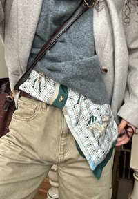 Μάλλινο φουλάρι με καρώ σχέδιο και πράσινες λεπτομέρειες, που απεικονίζει ένα άλογο. Φορεμένο κάτω από ένα γκρι πουλόβερ και στρωμένο κάτω από ένα μπεζ παλτό.