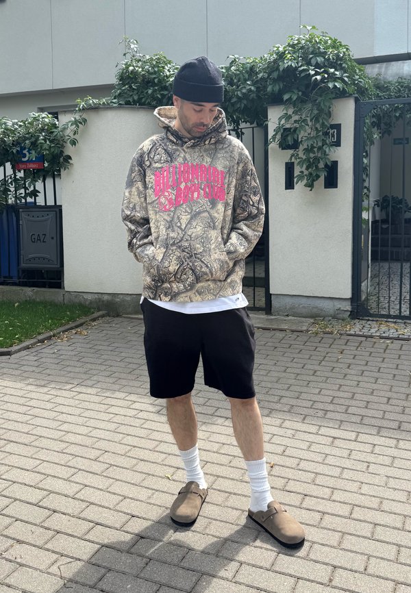Mann in Camouflage-Hoodie mit dem Text "Billionaire Boys Club", schwarzen Shorts, weißen Socken, braunen Slip-on-Schuhen und schwarzer Mütze, der auf dem Gehsteig steht.